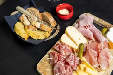 Gros plan sur une table basse avec une main tenant un verre de vin, accompagnée d’une planche apéritive de fromages et charcuterie.
