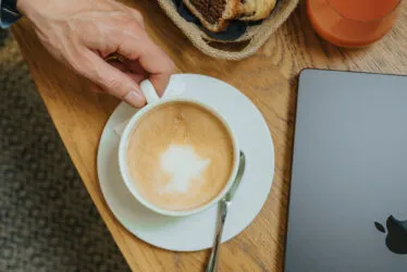Gros plan sur une table basse avec vue sur un café crémeux, un panier de viennoiseries et un ordinateur portable refermé.