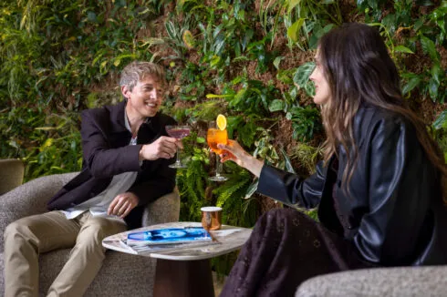 Couple seated in the hotel's designer armchairs, toasting with cocktails in front of a lush green wall.