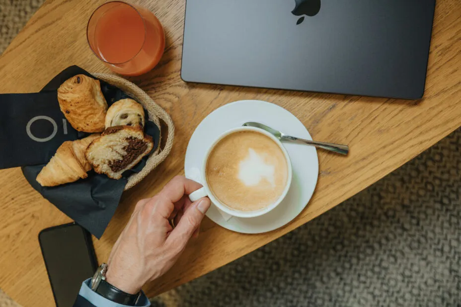 Top-down close-up: a coffee table with a creamy coffee, a basket of pastries, a fruit juice and a closed laptop.