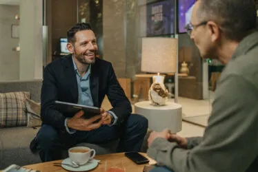Professionnel souriant assis sur le canapé de l’hôtel, tenant une tablette et discutant avec un interlocuteur vu de dos.