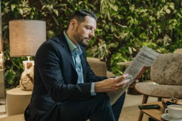 Homme d'affaire lisant un journal confortablement assis dans un salon de l'hôtel, avec une lampe allumée et le mur végétal en arrière-plan.