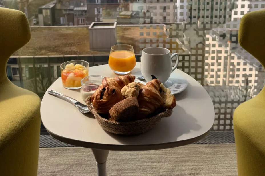 Gros plan sur café, viennoiseries et jus d’orange sur la table du coin salon en bow window, avec vue plongeante sur la ville.