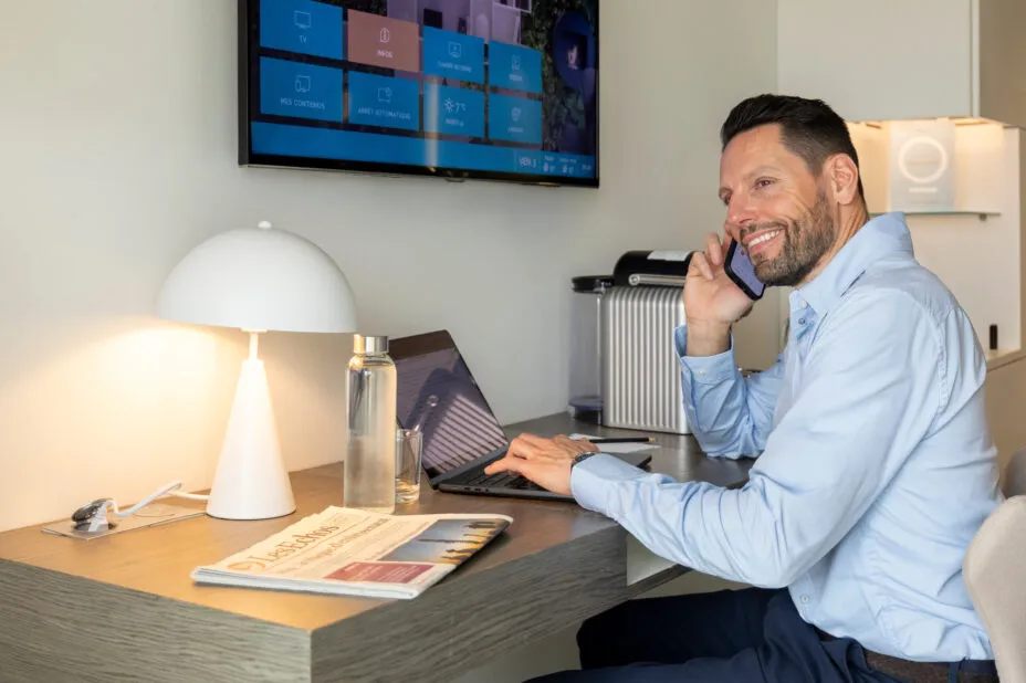 Homme d’affaires souriant assis au bureau de sa chambre de l’hôtel, au téléphone, avec journal et ordinateur portable sur le bureau.