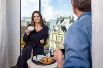 Couple complice savourant café et viennoiseries dans le salon bow-window de leur chambre de l’hôtel, ambiance lumineuse et détendue.