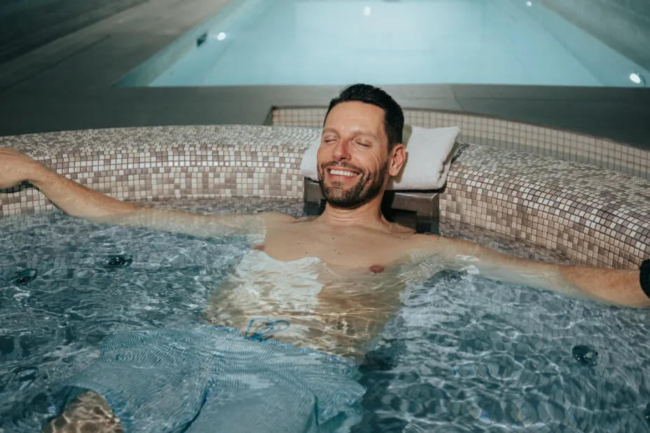 Homme détendu et souriant dans le jacuzzi de l’hôtel, avec la piscine en arrière-plan.