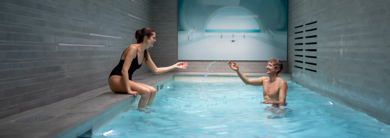 Couple having fun in the Saint-Antoine pool. The woman sits on the edge, splashing water at the man in the pool.
