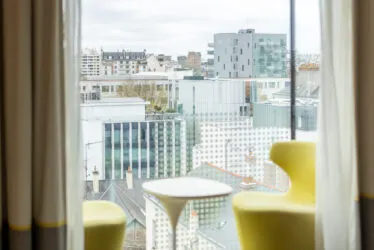 Urban view from the hotel window with a modern yellow armchair in the foreground and contemporary buildings in the background.