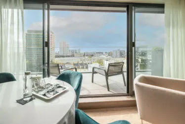 Vue dégagée sur la ville depuis la suite Saint-Antoine avec fauteuils et baie vitrée donnant sur un panorama urbain lumineux.
