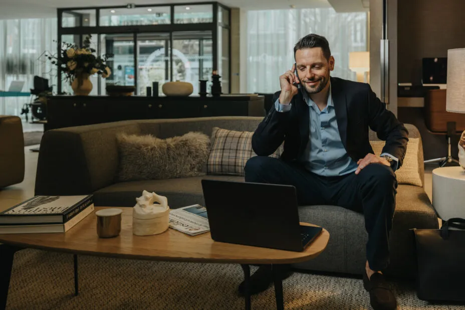 Hotel lounge area with a man on the phone seated on a sofa, laptop on the coffee table in a modern, bright setting.