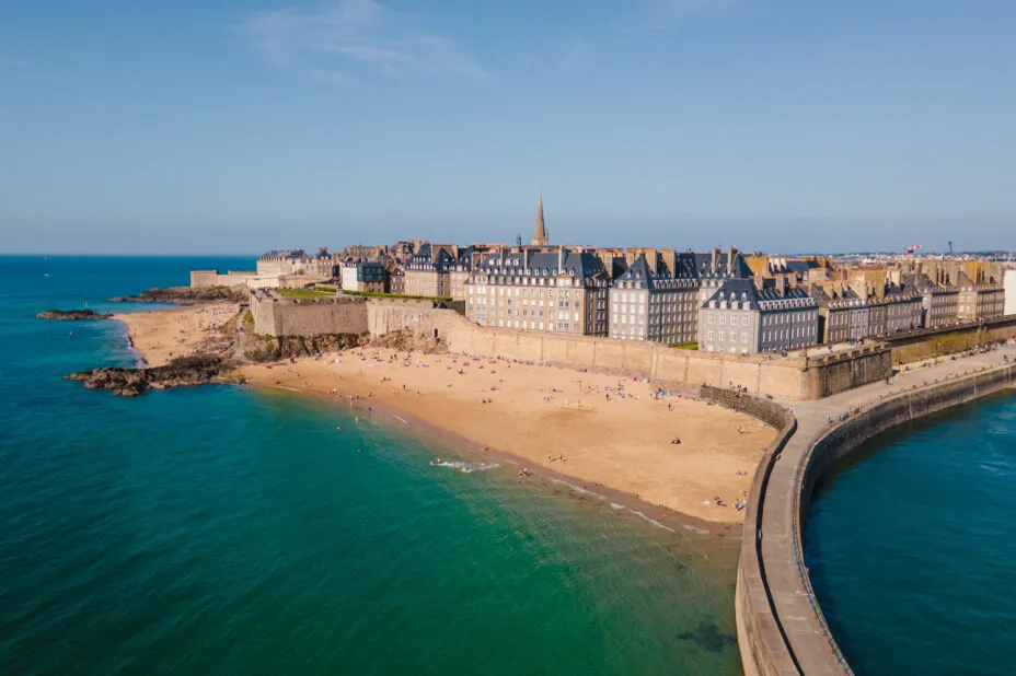 Vue aérienne de Saint-Malo sous le soleil avec plage au pied des remparts, et jetée protégeant le port.