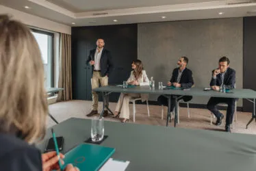 Seminar in progress in a hotel meeting room with a host and attentive participants.