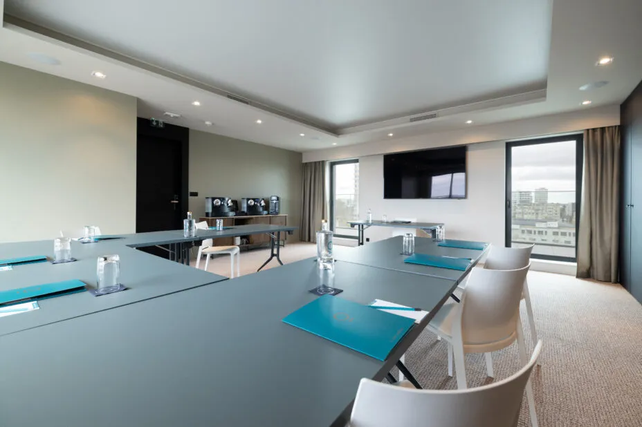 Perspective de la salle de séminaire de l'hotel, avec vue sur les tables, le grand écran de présentation et le coin café.