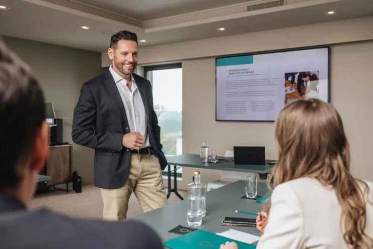 Seminar at Saint-Antoine: a smiling host standing and speaking with a participant seen from behind, with a presentation screen in the background.